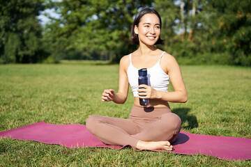 Wall Mural - Smiling fitness girl sits on mat and drinks water after workout, finish yoga training in park, exercise on fresh air