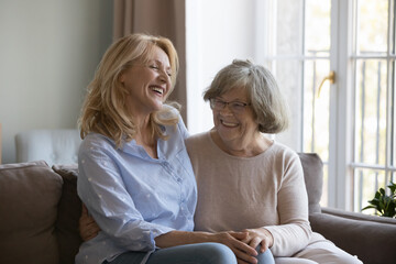 Laughing middle-aged woman enjoy pleasant positive talk with older mother sit on couch at home, happy multi generational relatives female joking, having friendly conversation appreciate time together