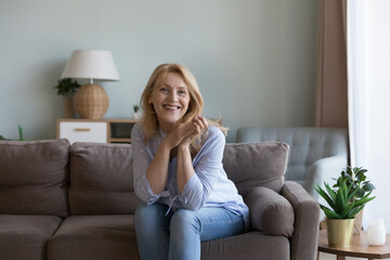 Pretty middle-aged woman posing for picture seated on sofa in modern living room. Portrait of happy, good-looking optimistic female spend carefree leisure at home, enjoy untroubled life on retirement