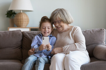 Pretty little 6s girl having fun using smart phone, making selfie picture, use new filter or mobile apps sitting on sofa with old great-grandmother enjoy modern tech usage and time together at home