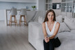 © Iona - Puzzled young beautiful caucasian woman in white blouse and black pants sits on cozy sofa home against kitchen. Tired blonde female in troubles looks at camera with thoughtful face expression at home.