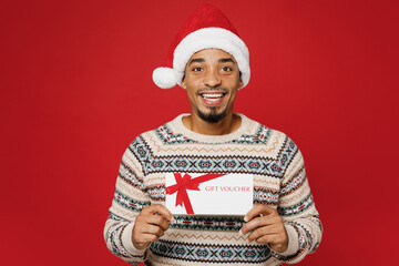 Sticker - Merry young man wear warm cozy Christmas sweater Santa hat posing hold gift certificate coupon voucher card for store isolated on plain red background. Happy New Year 2023 celebration holiday concept.