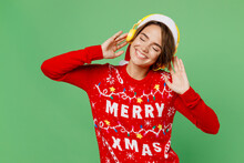 Merry Young Woman Wear Knitted Xmas Sweater Santa Hat Headphones Posing Listen To Music Close Eyes Enjoy Have Fun Isolated On Plain Pastel Light Green Background. Happy New Year 2023 Holiday Concept.