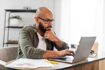 Portrait of concentrated male employee working on laptop at desk at home office, checking online project, copy space
