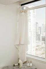 Wall Mural - A wedding dress, shoes and a bouquet of the bride are hung by the window in a hotel room against the backdrop of urban development. Wedding accessories of the bride on the wedding day and preparation