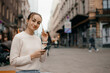 Young stylish woman in sweater using a smart phone standing outdoors on the street in Warsaw. Portrait of beautiful woman reading message on the street.