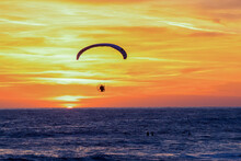 Solo Parasailer Paramotoring Over The Ocean At Sunset With Surfers In The Water.