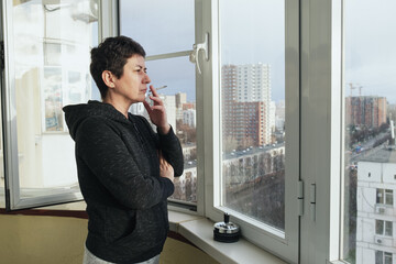 Middle-aged brunette woman stands on the glazed balcony of her apartment and smokes. A woman with a short haircut smokes a cigarette. Prohibition of smoking in residential premises.