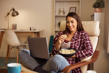 Happy woman eats popcorn while binge watching on laptop at home.