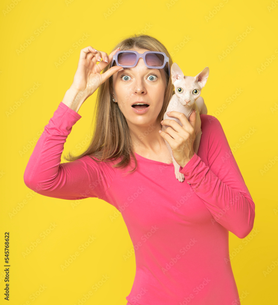 Beautiful surprised woman in red dress taking off glasses holding sphynx cat on shoulder over yellow background. Look at camera. shocked woman holding eyelets peek out glasses with wide open eyes