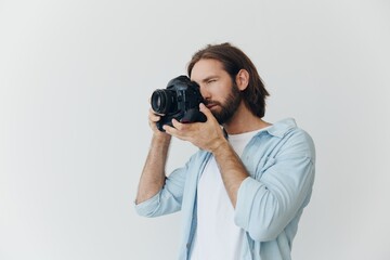 Poster - A male hipster photographer in a studio against a white background looks through the camera viewfinder and shoots shots with natural light from the window. Lifestyle work as a freelance photographer