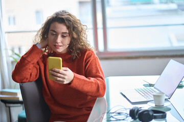 Young adult pretty woman holding smartphone using cellphone apps modern technology, looking at mobile phone while remote working or e learning from home sitting at table with laptop computer.