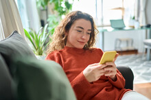 Smiling Relaxed Young Woman Sitting On Couch Using Cell Phone Technology, Happy Lady Holding Smartphone, Scrolling, Looking At Cellphone Enjoying Doing Online Ecommerce Shopping In Mobile Apps.