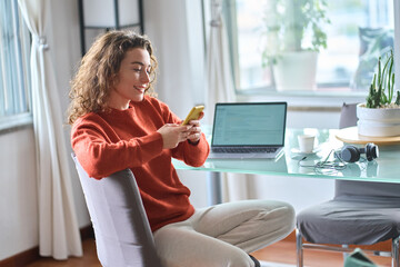 Young smiling pretty woman holding smartphone using cellphone modern technology, looking at mobile phone while remote working or e learning from home sitting at table with laptop computer.