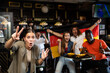 © JackF - Cheerful excited young female fan watching sports game in bar, gesturing emotionally, happy with victory of favorite team against background of people with national flag of Germany..