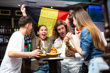 Wall Mural - Excited diverse soccer fans with flag of Belgium celebrating victory of team with pint of beer and chips in the pub