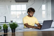 © F8  \ Suport Ukraine - Positive indian man looking for job online, sitting at table, using laptop and taking notes, kitchen interior, copy space. Excited indian man student working online from home, remote job