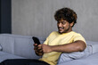 © F8  \ Suport Ukraine - Young indian man with headphones on his neck holding phone and sitting on the sofa at home
