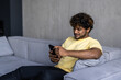 © F8  \ Suport Ukraine - Young indian man with headphones on his neck holding phone and sitting on the sofa at home