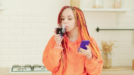 Wall Mural - Young woman with bright long dreadlocks browsing mobile phone, drinking red wine, dancing in kitchen.