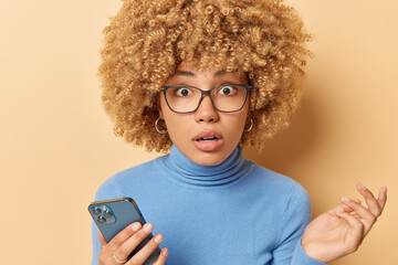 Poster - Portrait of curly haired woman stares through eyeglasses holds mobile phone reacts to shocking news sends text messages wears blue turtleneck isolated over beige background. Human reactions.