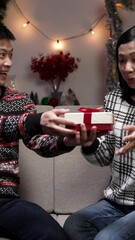 Wall Mural - Vertical Screen: cheerful asian male surprising his excited wife by taking out a gift box while they are sitting on the sofa in a cozy Christmas living room at home in the evening