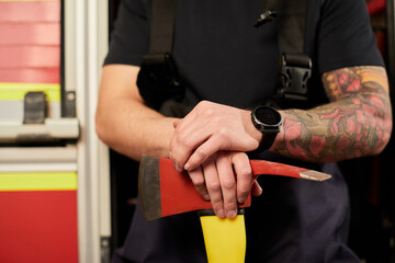 Young handsome adult, muscular firefighter in uniform holding ax fire equipment in hands, isolated on truck background. Portrait strong fireman in fireproof uniform holding an ax in his hands