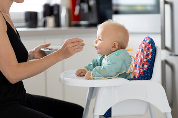 Wall Mural - Mother spoon feeding her baby boy child in baby chair with fruit puree in kitchen at home. Baby solid food introduction concept.