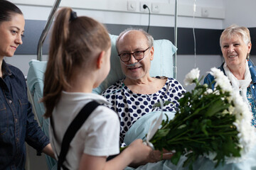 Wall Mural - Sick grandfather receiving flowers from granddaughter in hospital room. Bedridden old man being visited by relatives in geriatric clinic room. Little girl giving flowers to bedridden elderly man.