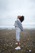 Fitness woman working out and stretching at the beach towards under autumn rainy weather. Afro hairstyle sporty black female doing breathing exercise outdoor.