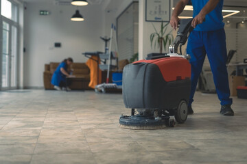 Cleaner cleans hard floor with scrubber machine while other cleaner cleans in the background