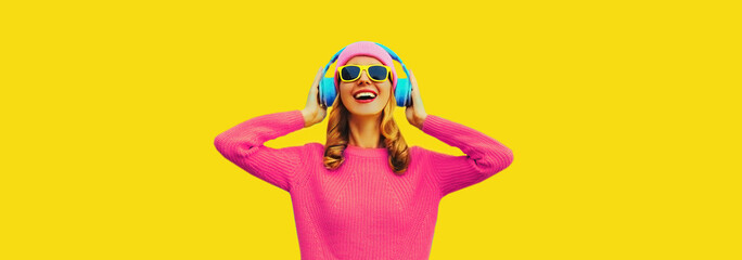 Wall Mural - Portrait of happy smiling young woman with headphones listening to music wearing colorful pink sweater on yellow background