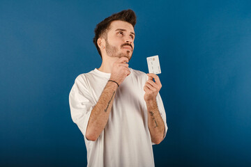 Wall Mural - Caucasian young man wearing t-shirt posing isolated over blue background holding credit or debit card serious face thinking about question, very confused ide