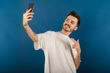 Wall Mural - Handsome young man wearing casual clothes posing isolated over blue background pointing index finger on mobile phone while taking selfie with big smiles. Communicating concepts.