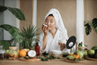 © sofiko14 - Young asian woman in white bathrobe and towel holding half of coconut and making homemade cream for healthy skin. A lot of ingredients for homemade cosmetics lying on wooden table.
