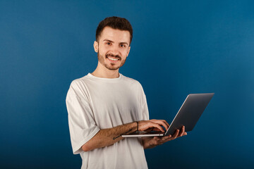 Wall Mural - Happy young man wearing white t-shirt posing isolated over blue background looking at the camera and using computer laptop smiling with a happy and cool smile on face. Showing teeth.