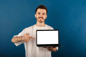 Wall Mural - Cheerful young man wearing t-shirt posing isolated over blue background pointing with index finger to laptop pc computer with blank empty screen