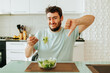 A bowl of salad is on the kitchen table, a young man took a fork and is preparing to eat a salad, showing a dislike and making a grimace of displeasure. Nobody likes healthy food.