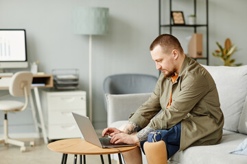 Side view portrait of man with prosthetic leg using laptop while working in minimal office interior, copy space