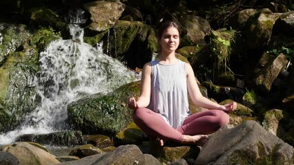 Sticker - Beautiful young woman meditating near waterfall in mountains on sunny day