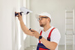 © Studio Romantic - A Caucasian male repairman in uniform and glasses holds a screwdriver and installs video surveillance equipment on the wall inside the room. The concept of security and protection.