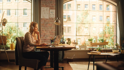 Aufkleber - Young Pretty Woman with Blond Hair Wearing Headphones and Working from Home on Laptop Computer in Stylish Loft Apartment. Creative Female Smiling, Checking Social Media, Browsing Internet.
