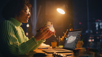 Leinwandbilder - Concentrated Black Woman Draws on Shoe and Looks at Laptop, Working at Home in the Evening. Individual Drawing on Sneakers Concept. Work at Night In Loft Apartment Living Room.