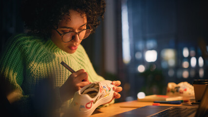Leinwandbilder - Female Designer Puts Patterns on Shoe and Comparing with 3D Model on the Laptop Screen. Black Woman Feel Inspired while Drawing in the Evening. Creating Trendy Sneakers Concept.