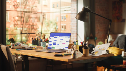 Poster - Shot of a Laptop Computer with Openned Calendar App Standing on the Wooden Desk in the Creative Cozy Living Room. In the Background Sun is Shining Through the Window in Open Space Studio.
