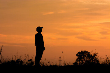 Wall Mural - a silhouette of a person in a cap standing in tall grass, with a sunset in the background.