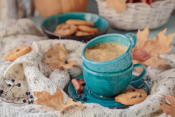 Wall Mural - Autumnal composition with white knitted scarf, blue cup of coffee, cookies and dry yellow leaves on a table. Autumn mood.