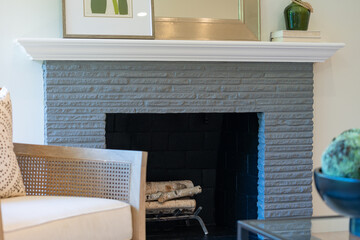 Old fashioned painted brick fireplace with birch logs, white wood mantlepiece and neutral rattan cushioned side chair.