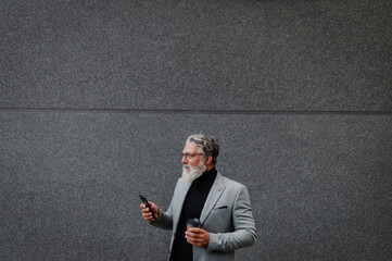 Senior business man standing outside while using smartphone and drinking coffee