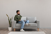 Handsome bearded man using laptop on grey sofa near light wall